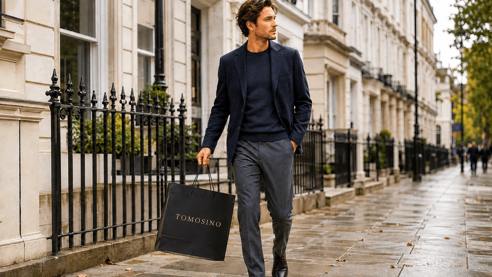 A man in a navy blazer and knit walking a rain-washed London street, carrying a black Tomosino shopping bag.
